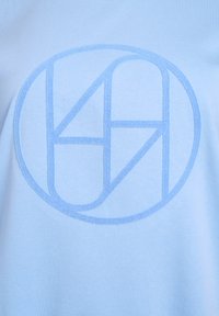 Light blue fabric with a soft texture featuring a circular logo in a darker blue, incorporating abstract shapes and the number 74.