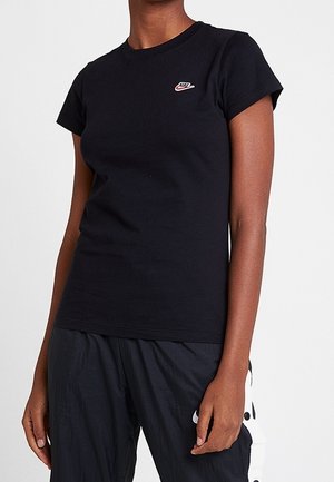 Persoon draagt een zwart Nike T-shirt en zwarte trainingsbroek met witte zijkanten, staand voor een effen lichte achtergrond.