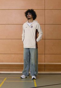 Beige fleece trui met bruine accenten, voorzien van een halve rits en logo. Gecombineerd met losse blauwe jeans en witte sneakers.