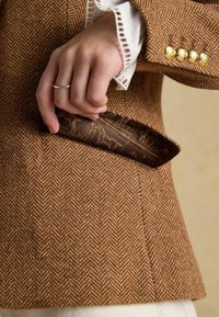 Sacou maro cu model herringbone și nasturi aurii, având o mână care ține un obiect maro cu model parțial vizibil în buzunar.