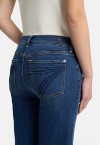 Ciemnoniebieskie jeansy z dżinsu o dopasowanym kroju, z tylnymi kieszeniami ozdobionymi przeszyciami oraz małą metką w pobliżu talii.