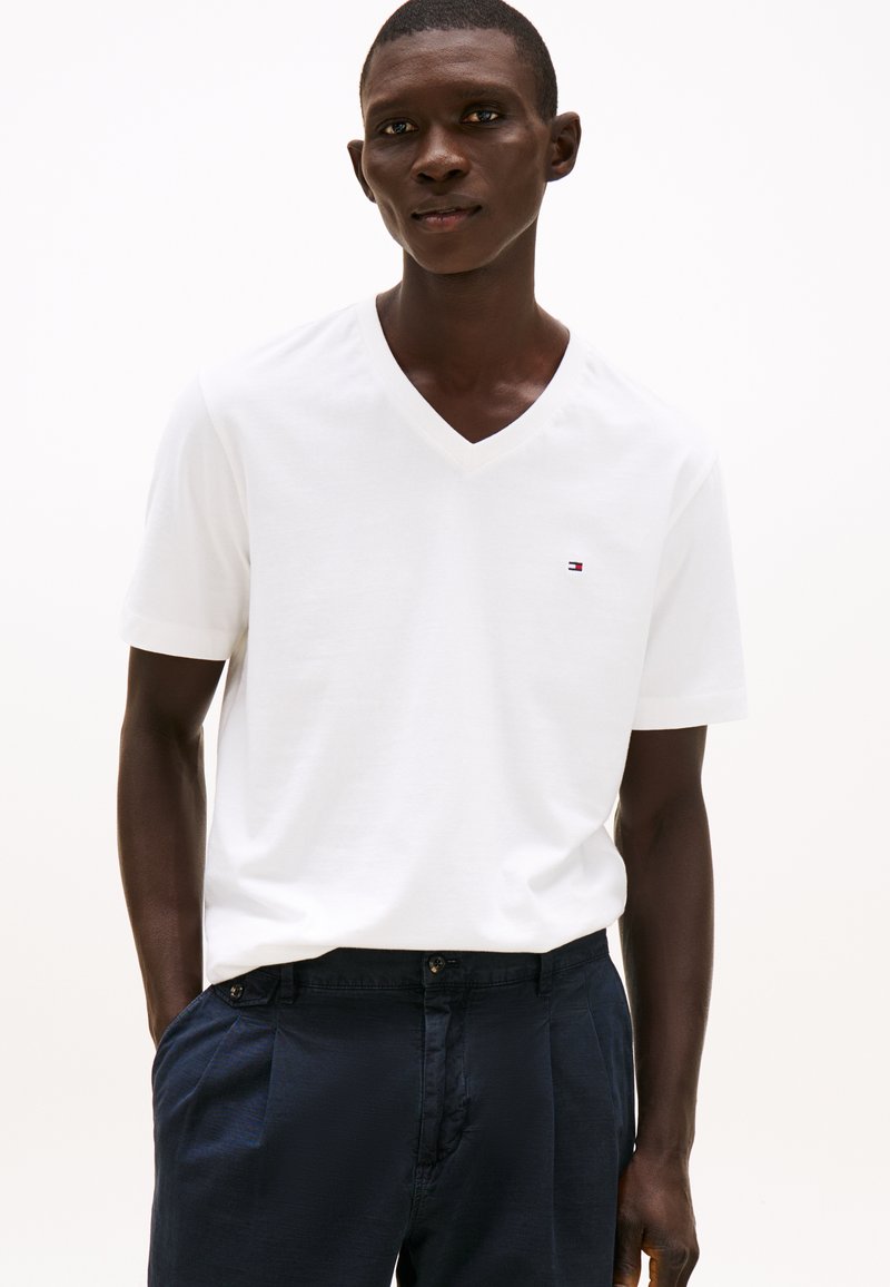 White cotton V-neck t-shirt with short sleeves, featuring a small logo on the left chest. Worn with dark navy trousers.