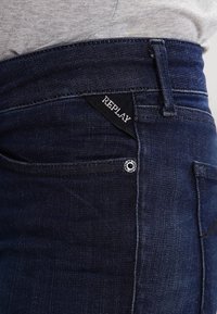 Jeans de mezclilla azul oscuro con textura tejida, que presentan una etiqueta triangular negra de Replay y un acento de botón plateado en el bolsillo frontal.