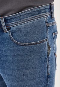 Blaue Jeans aus Denim mit geradem Schnitt, ausgestattet mit orangefarbenen Nähten, fünf Taschen und einem schwarzen Stoffetikett auf der Gesäßtasche.