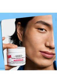Kiehl's Ultra Facial Cream-krukke, hvit med rød og svart tekst. Nærbilde av et ansikt med glatt hud og langt mørkt hår, som holder krukken.