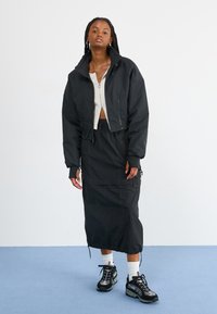 Jachetă scurtă neagră cu guler înalt, asortată cu o fustă lungă neagră. Material texturat și pantofi sport negri cu accente reflectorizante.