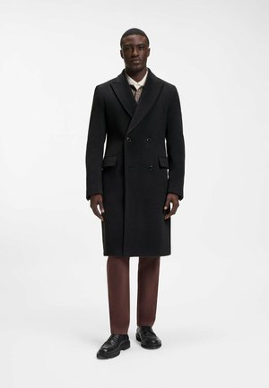 Man draagt een zwarte dubbelgeknoopte wollen jas, bruine broek, zwarte schoenen en een geruit overhemd met een witte kraag, staand tegen een witte achtergrond.