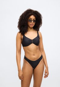 Černý bikiny set se strukturovaným podprsenkovým topem s ústřední přezkou a nízkým pasem kalhotek, oba vyrobené z hladkého, pružného materiálu.