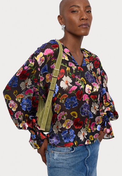 Blusa floreale con fiori multicolore vivaci su un tessuto nero trasparente. Presenta una scollatura a V, maniche lunghe e polsini arricciati. Indossata con una borsa verde.
