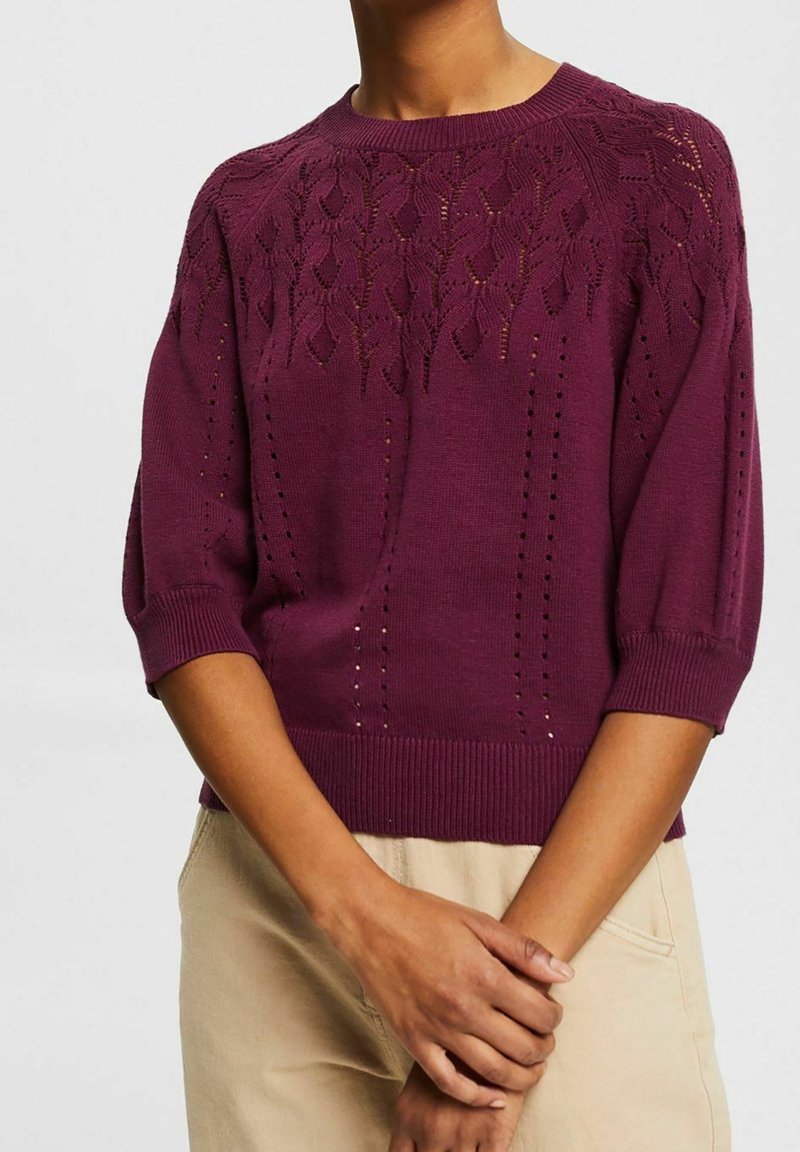 Femme portant un pull en maille prune avec un motif de feuille en dentelle près de l'encolure et des manches trois-quarts, associé à un pantalon beige clair.