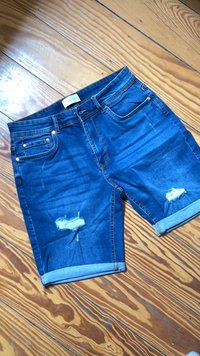 Pantalones cortos de mezclilla azul con puños doblados y parches desgastados en ambas piernas, colocados en el suelo de madera.