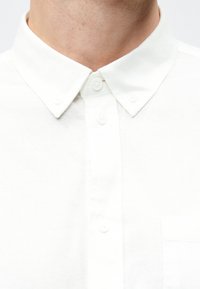 Weißes Hemd mit Knopfleiste und Kragen, mit einer Vorderbrusttasche, aus strukturiertem Stoff, Nahaufnahme des oberen Bereichs.