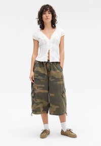 Blusa branca de mangas curtas com decote em babados, combinada com calções de camuflagem com bolsos laterais grandes e bainha ajustável com cordão.