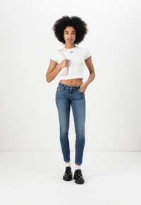 Fehér, rövidített póló kék logóval, sötét mosású skinny farmernadrág és fekete cipő. A modell egy világos rózsaszín táskát tart, és egy fehér háttér előtt áll.