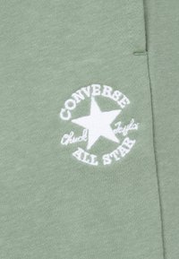 Grønt bomullsstoff har en hvit brodert logo med en stjerne og teksten "Converse Chuck Taylor All Star." Glatt tekstur, uformelt design.