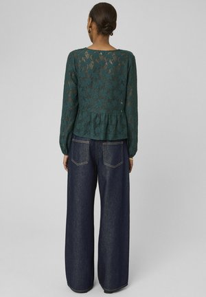 Femme avec les cheveux en chignon portant un chemisier en dentelle vert foncé à manches longues et un jean large bleu foncé, debout contre un fond gris, vue de dos.