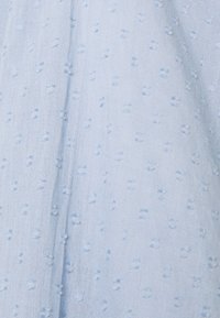 Tissu bleu clair avec des pois texturés, drapé doux et apparence légèrement translucide. Le matériau est léger et aéré.