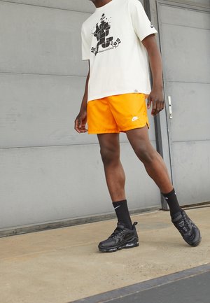 Vit grafisk t-shirt med en svart design, orange shorts, svarta Nike-sneakers och svarta strumpor, ståendes på en betongyta.