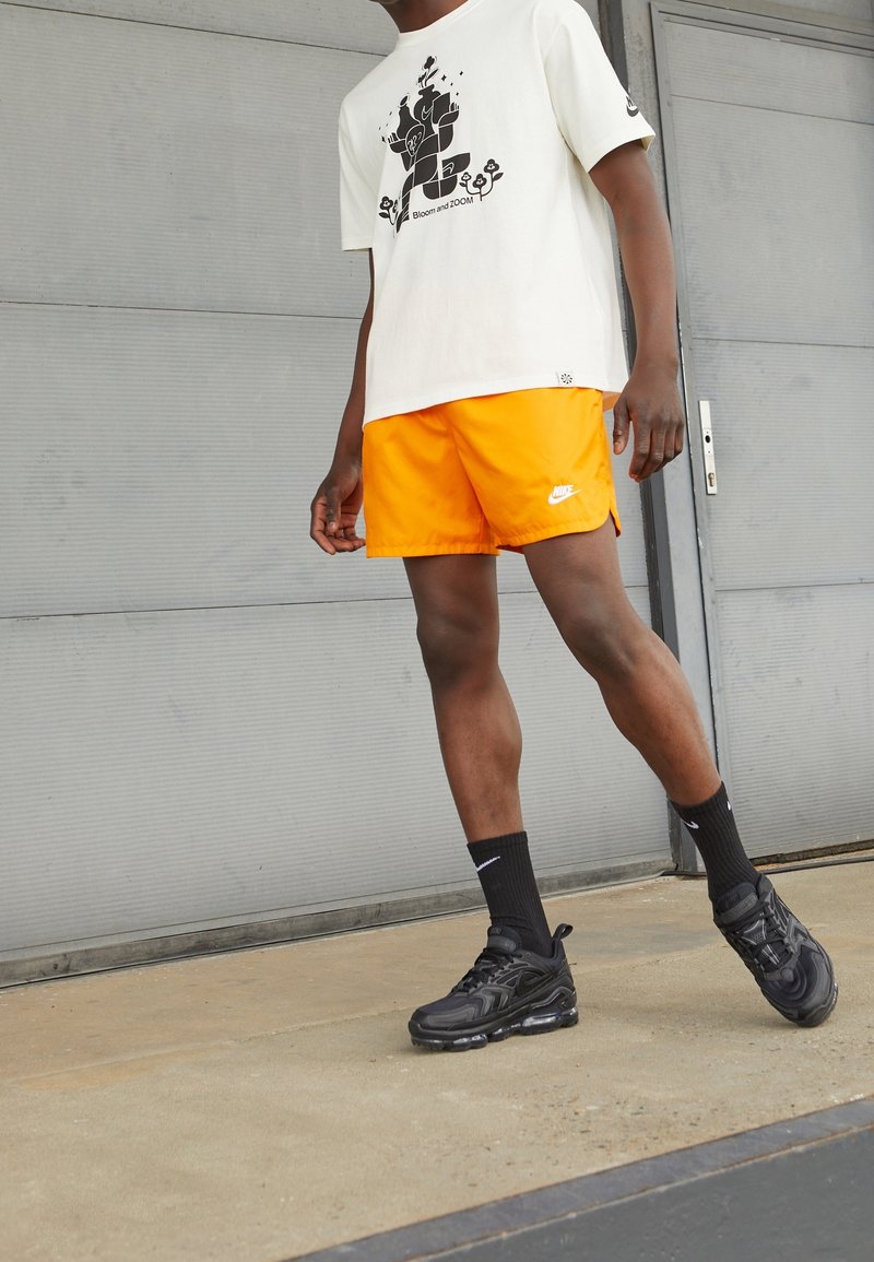 Biała koszulka graficzna z czarnym wzorem, pomarańczowe szorty, czarne sneakersy Nike i czarne skarpetki, stojąc na betonowej powierzchni.