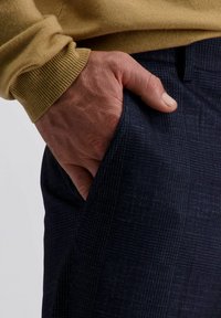 Hand in een donkerblauwe geruite broekzak met de mouw van een mosterdgele trui zichtbaar tegen een neutrale achtergrond.