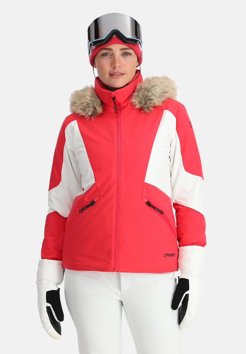 Heldere rode skijack met witte accenten en een met bont gevoerde capuchon. Beschikt over twee met rits afsluitbare zakken en wordt gedragen met witte broek en handschoenen.