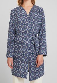 Blå mönstrad omslagsklänning med cirkulära mönster i nyanser av blått och vitt, långa ärmar och ett tygbälte i midjan.