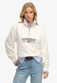 Kobieta ubrana w kremową bluzę z półzamkiem i napisem "SUPERDRY New York ATHL DEPT" oraz amerykańską flagą, nałożoną na niebieską koszulę w paski i dżinsy.