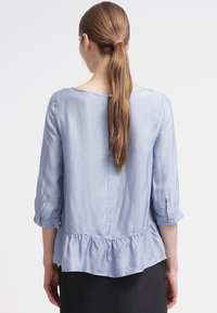 Ljusblå peplumblus i ett mjukt material med trekvartslånga ärmar, prydd med knappdetaljer och en samlad nederkant. Bakvy visas.