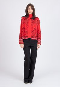 Röd mönstrad jacka med svarta accenter, med hög halsringning och långa ärmar. I kombination med svarta byxor och svarta skor.