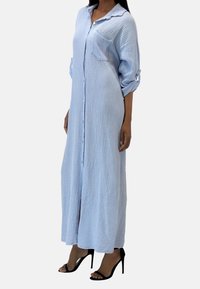 Robe en coton longue, de couleur bleu clair, avec un design texturé, fermeture à boutons et manches roulées. Présente une poche poitrine et des fentes sur les côtés.