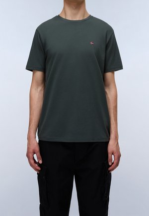 Homme portant un t-shirt manches courtes vert foncé avec un petit logo drapeau sur la poitrine et un pantalon cargo noir, debout devant un fond clair uni.
