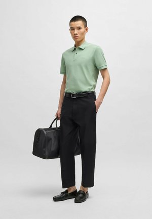Jasnozielona koszulka polo, czarne spodnie, czarny pasek i czarne loafersy. Nosi czarną torbę o uporządkowanym fasonie. Neutralne tło.