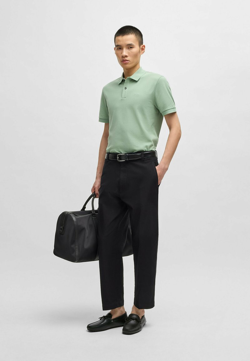 Jasnozielona koszulka polo, czarne spodnie, czarny pasek i czarne loafersy. Nosi czarną torbę o uporządkowanym fasonie. Neutralne tło.