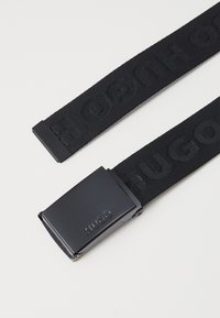 Fekete szövött anyagú öv matt fekete fém csattal. A csattón gravírozott logó található. Az övszíj "HUGO" feliratozása diszkrét mintázatban jelenik meg.