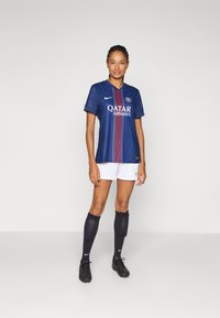 Nike women's soccer jersey in navy, featuring a prominent red and blue diamond pattern, white shorts, black socks, and cleats.