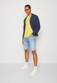 Cardigan de felpa azul marino, camiseta amarilla, pantalones cortos de mezclilla azul claro y zapatillas blancas. Los accesorios incluyen gafas de sol amarillas.