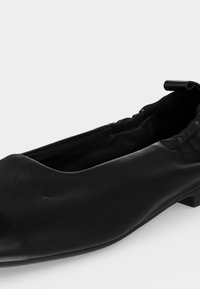 Μαύρο δερμάτινο επίπεδο παπούτσι με συγκεντρωμένη ελαστική λεπτομέρεια γύρω από τη φτέρνα και μια μικρή διπλωμένη δερμάτινη ταμπέλα στο πίσω μέρος.