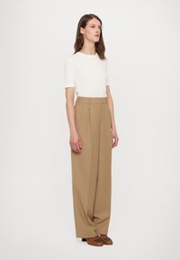 Haut blanc à manches courtes associé à un pantalon beige à taille haute et jambes larges, avec une texture lisse et des plis subtils. Chaussures à talons brunes.