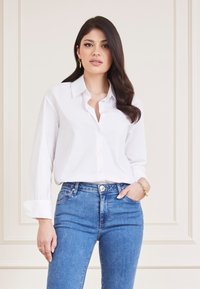 Camicia bianca con bottoni, maniche lunghe arrotolate, abbinata a jeans in denim blu. La camicia presenta un colletto classico e una vestibilità rilassata.