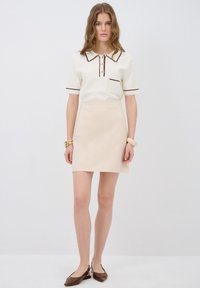 Haut blanc à manches courtes avec des bordures marron et une poche, associé à une jupe mini beige. Le mannequin porte des ballerines marron et des accessoires en or.