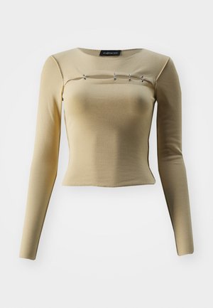 Langermet beige topp laget av myk stoff med en tettsittende design og rund hals, med tre dekorative sølvdetaljer over brystet.