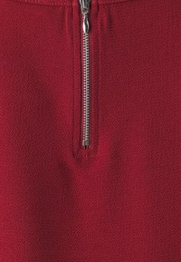 Detailní záběr na stříbrný zip na texturované tmavě červené tkanině, který ukazuje podrobný steh okolo oblasti zipu.