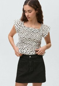 Haut blanc avec un motif tacheté noir, décolleté carré et manches courtes à volants. Associé à une jupe en denim noire avec des poches avant.
