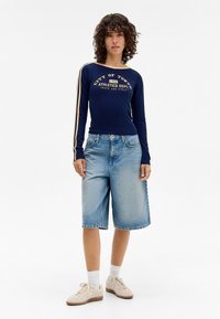 Marineblå langermet skjorte med gulltekst og kremfargede striper, kombinert med lyseblå knelange denimshorts og beige sneakers.