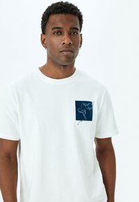 Camiseta de algodón blanca con cuello redondo y mangas cortas. Presenta un diseño de bolsillo cuadrado en navy con un detalle de dibujo a línea.