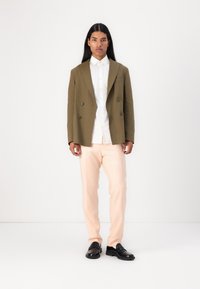 Olijfgroene dubbelknop blazer boven een wit shirt, gecombineerd met licht perzikkleurige broek en zwarte instappers, tegen een effen achtergrond.