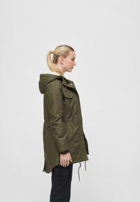 Olive green parka with a hood, featuring a zip and button closure, front pockets, and adjustable drawstring at the waist. Smooth texture.