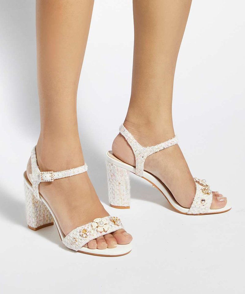 Pieds portant des sandales blanches à talon bloc texturé avec brides à la cheville et embellissements floraux sur les brides avant sur un fond uni.