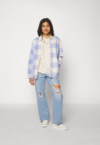 Overmaat, lichtblauw en wit geruite fleece cardigan, gecombineerd met een crèmekleurig shirt, versleten blauwe jeans en witte sneakers.