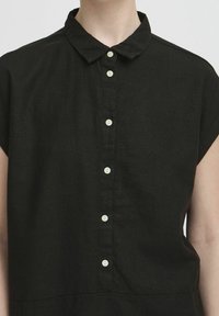 Camicia nera a maniche corte con colletto, caratterizzata da sette bottoni bianchi sulla parte anteriore, indossata da una persona.
